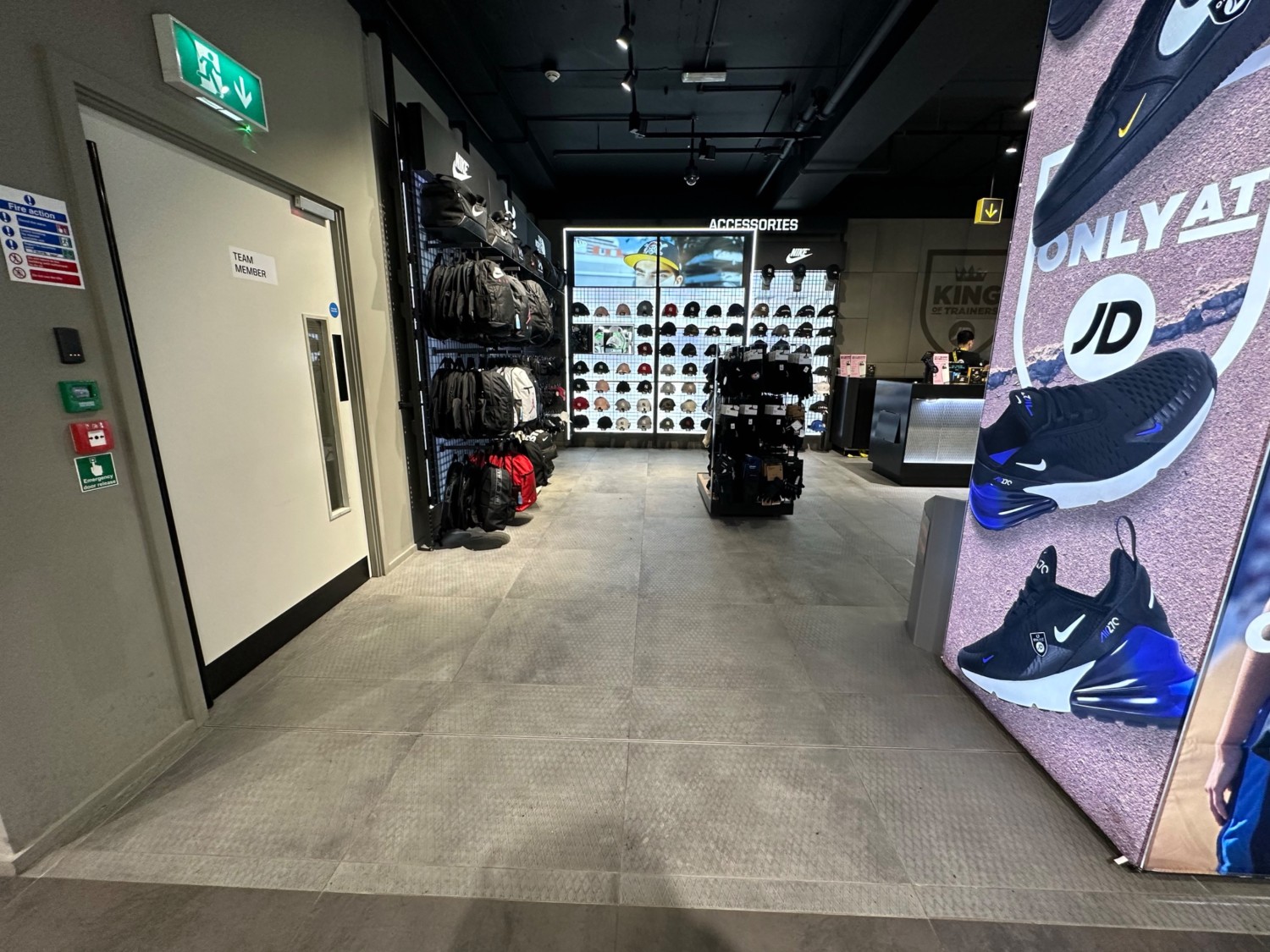 From end of path in far left corner facing straight. Third pillar on right. Path ahead leads to bags and hats display area along back wall ahead.
