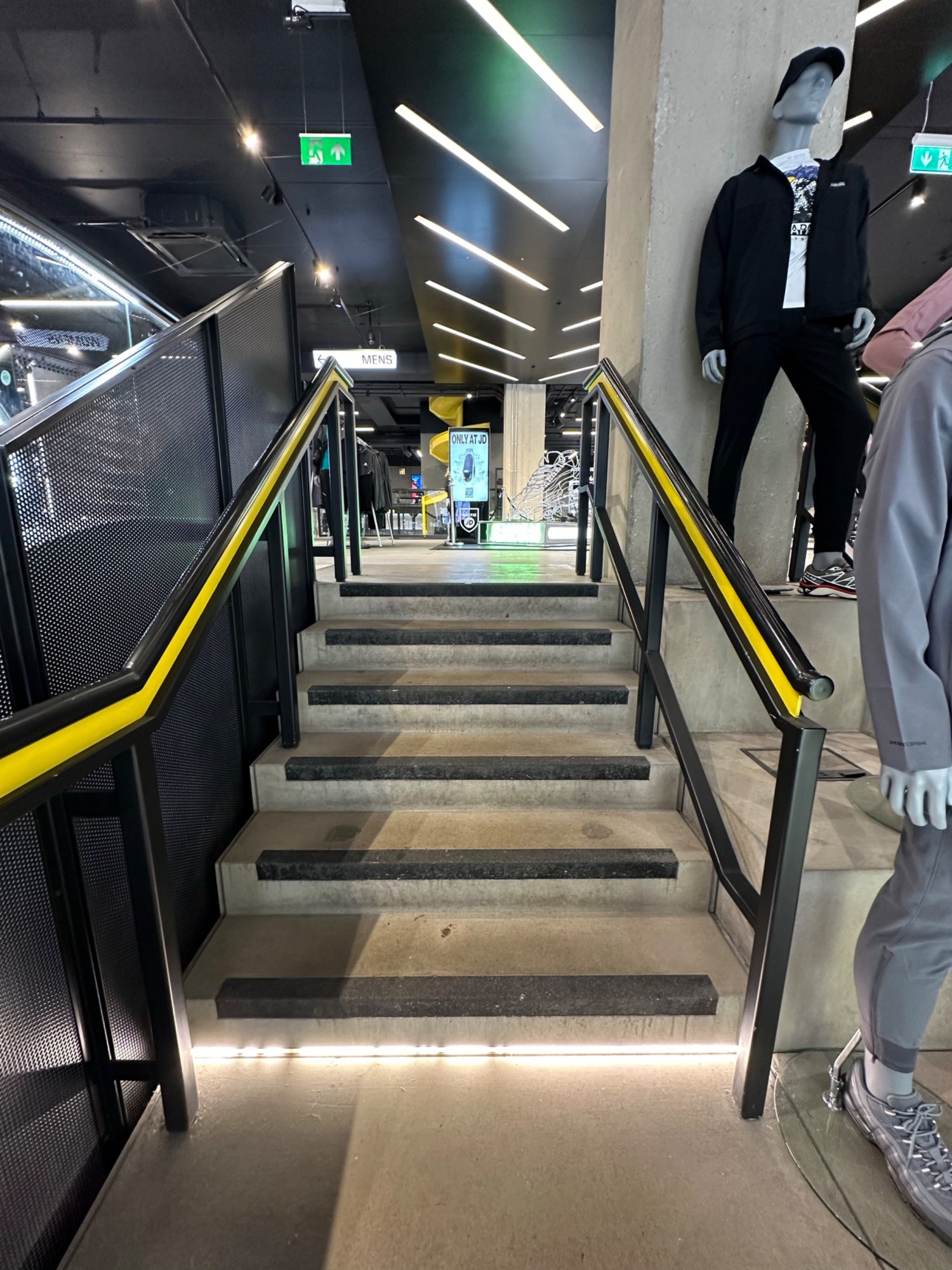 From top of second flight of stairs facing straight. Black and yellow handrail on left and right of third flight of stairs straight ahead.