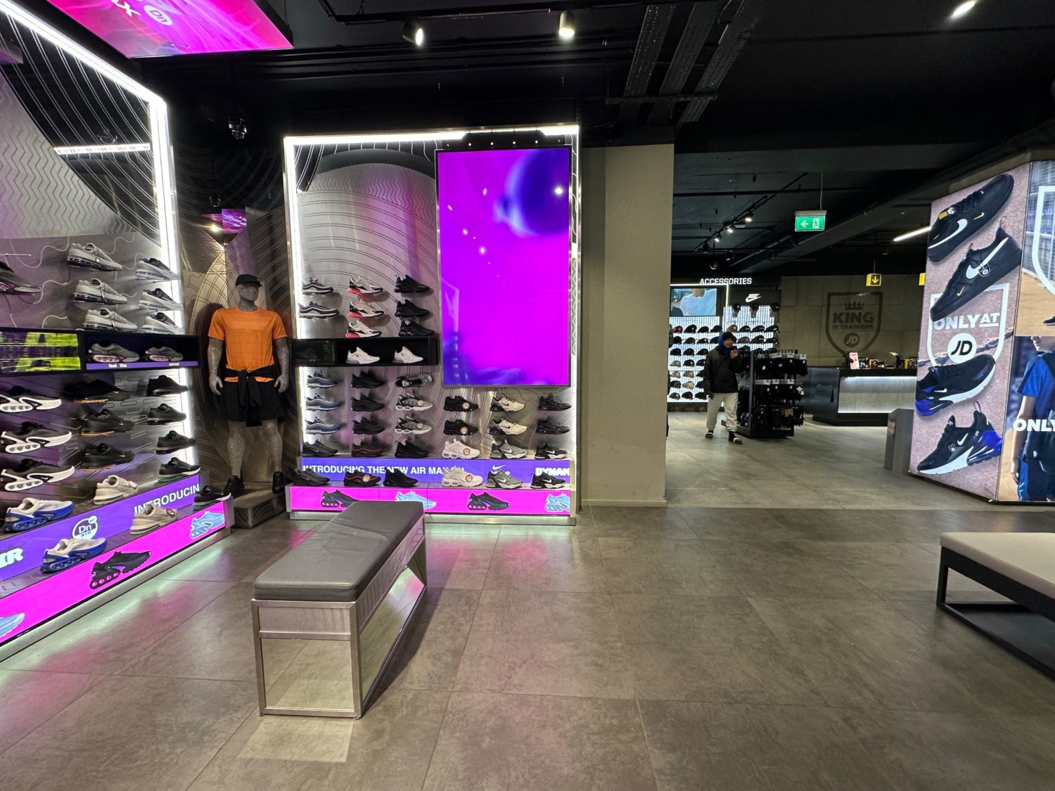 From end of central shoe display facing straight. Shoe wall to far left. Benches on left and right of path. Path leads to back left area to far right.