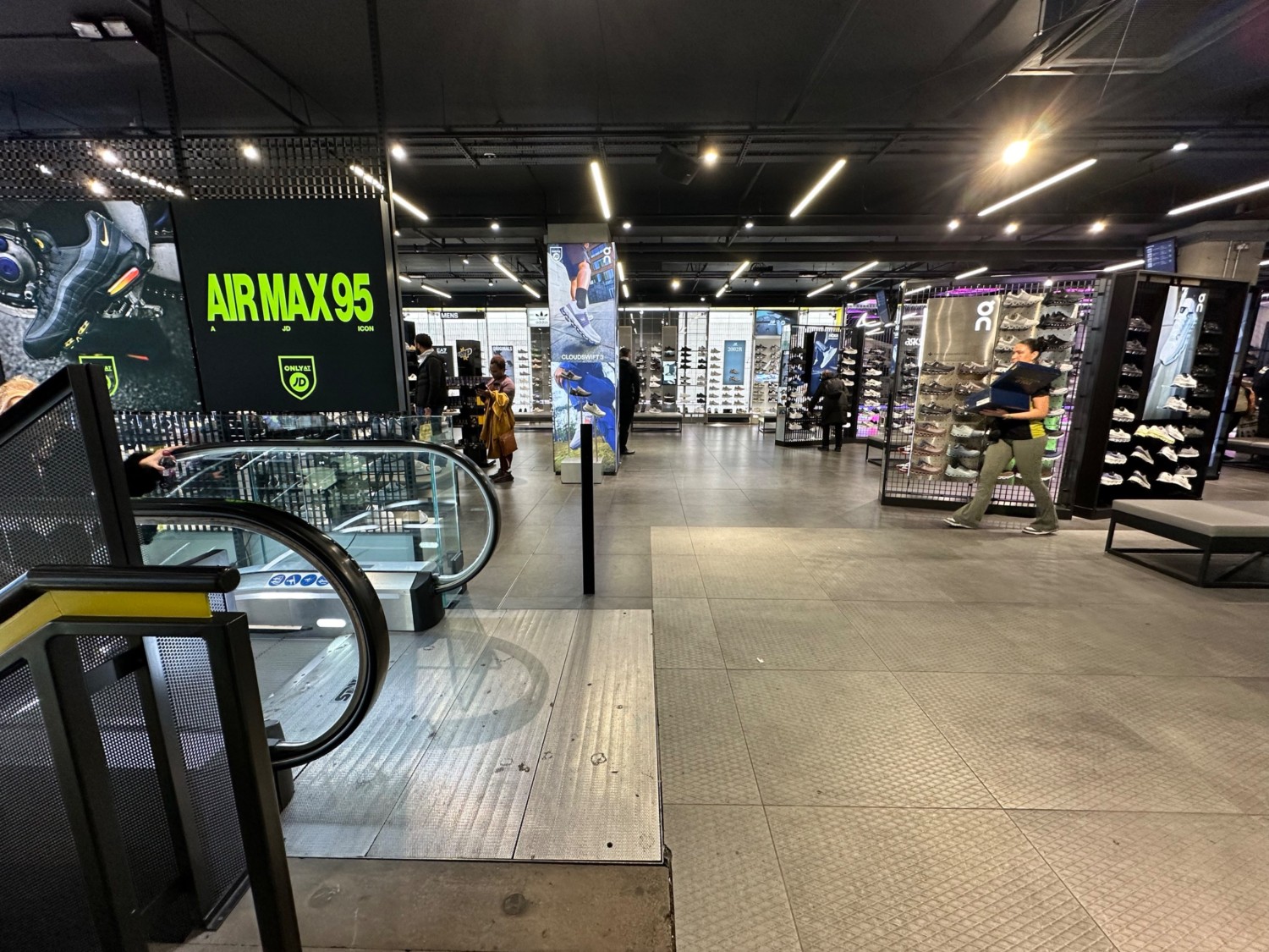 From top of stairs and escalator facing left. Bench and shoe rack on right. Bollard ahead past escalator on left.  Path ahead to shoe area.