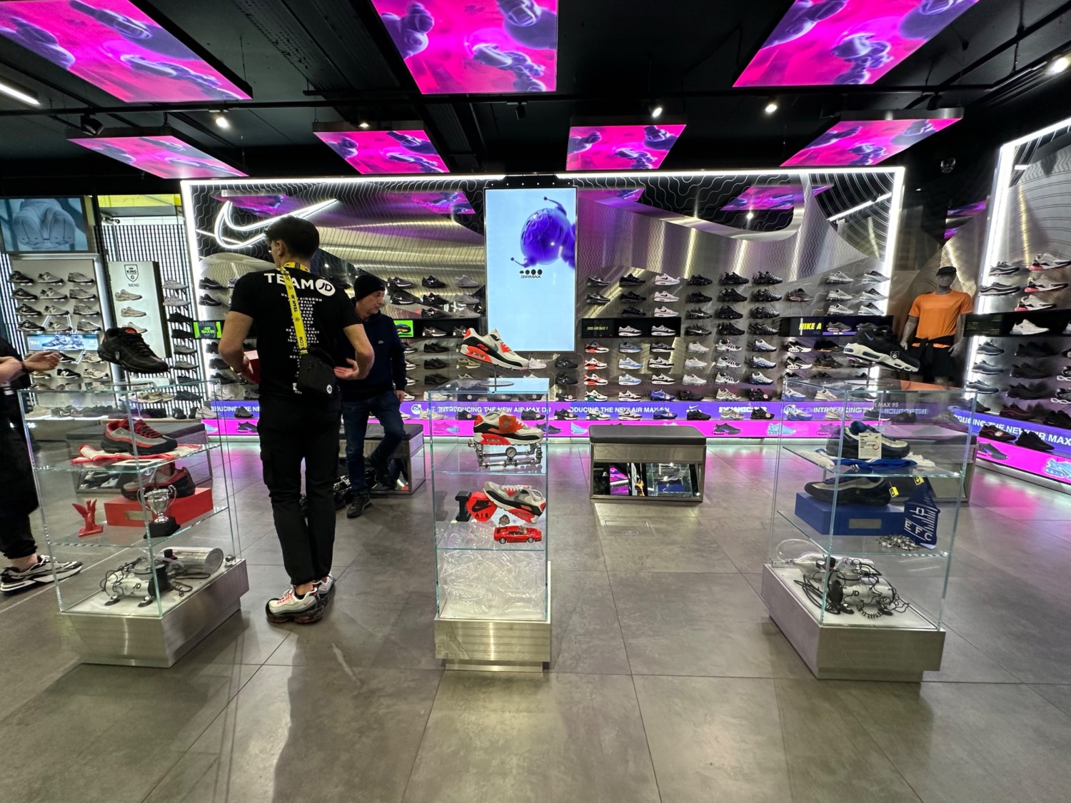 From central shoe display facing left. Glass display boxes spaces evenly out ahead. Benches spaced evenly out before shoe rack along back wall ahead.