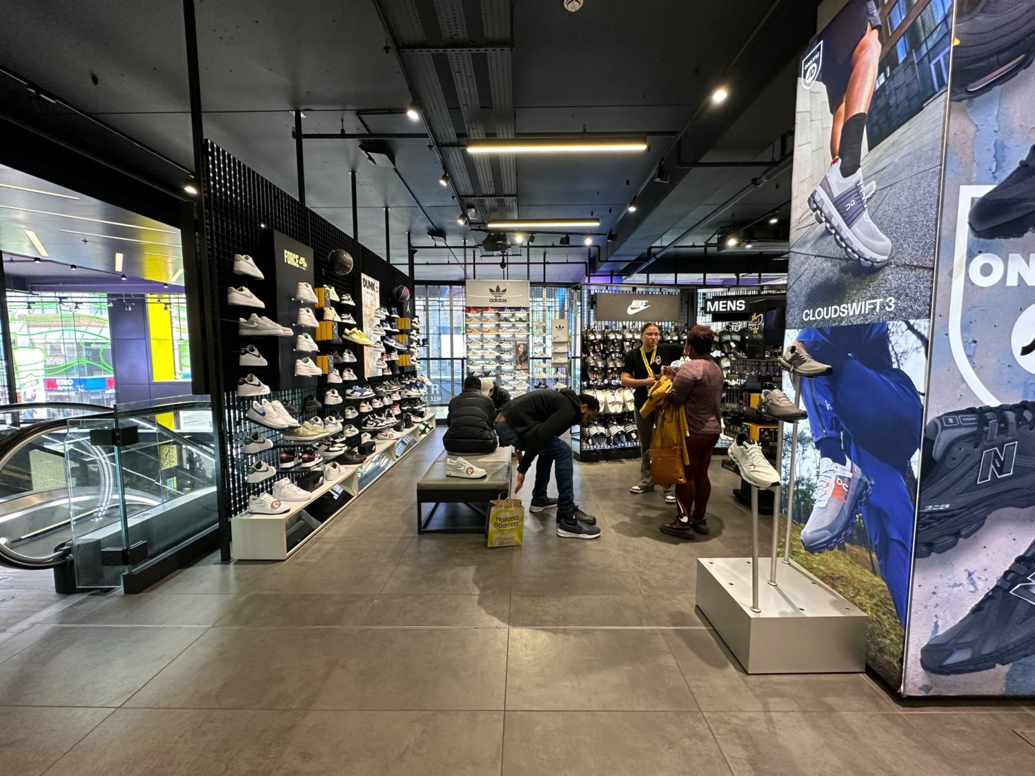 From left of escalator facing back. First pillar on right. Path ahead leads to front left area. Shoe displays along left and back. Bench in path ahea