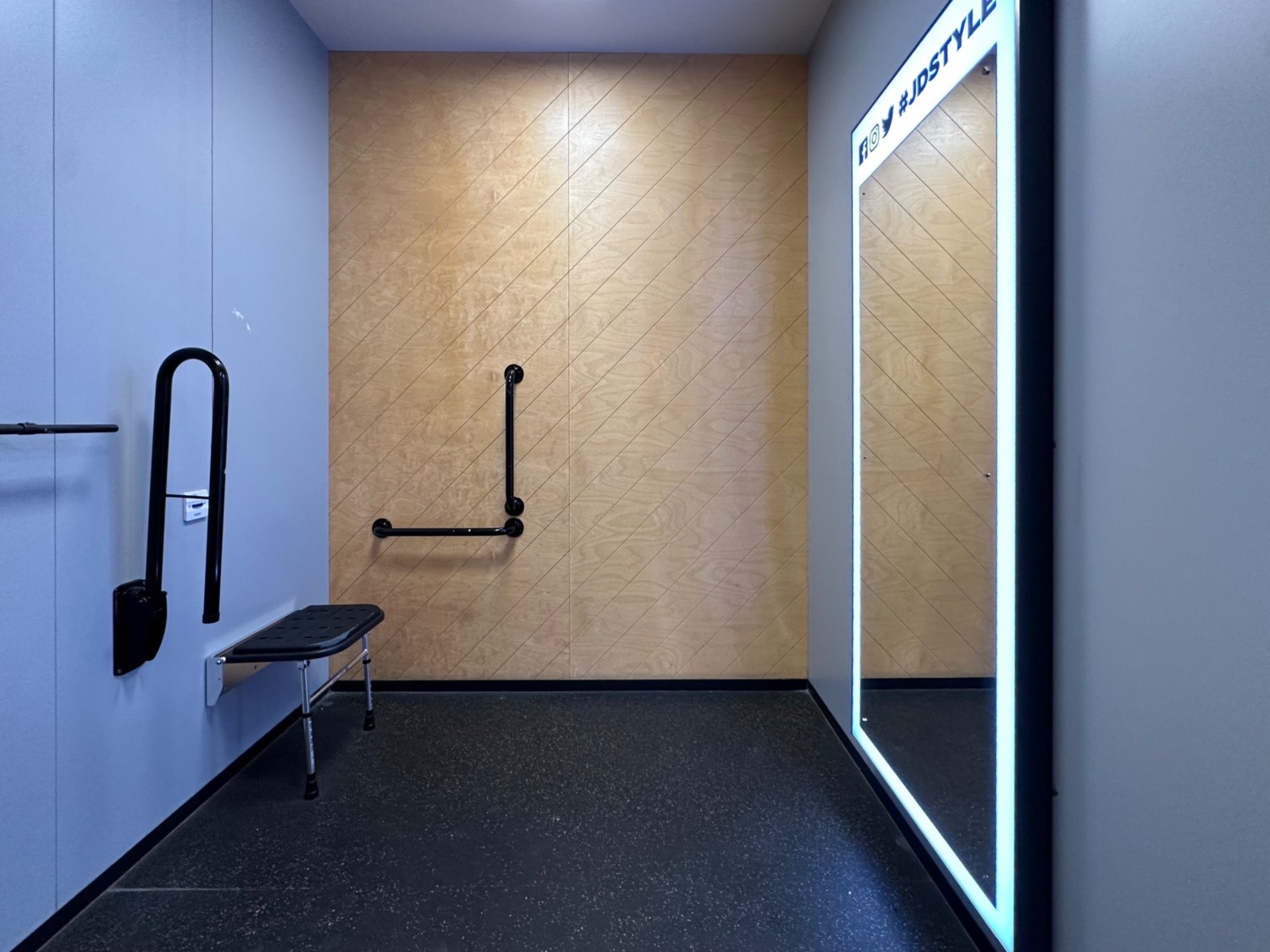 From door facing straight, bench on left end corner with grabrails on both sides. Full body mirror on right wall.