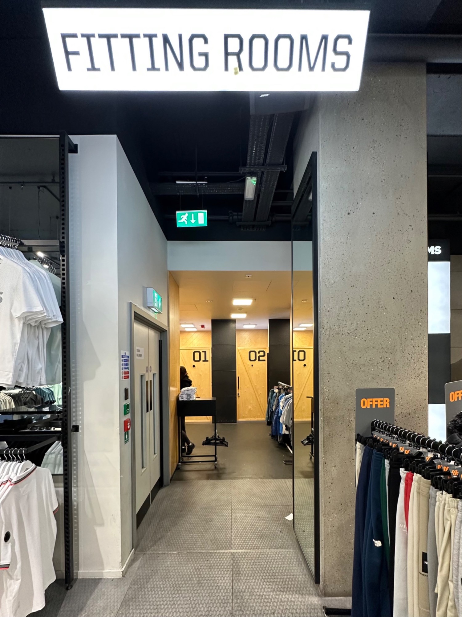 From start of path to left of payment area looking straight. Clothing racks to left and right followed by open entrance to fitting rooms.