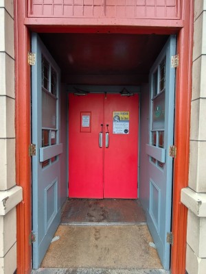 From end of outdoor path looking straight. Grey door leading to another set of doors. Push in centre to open. Carpet on threshold of door.