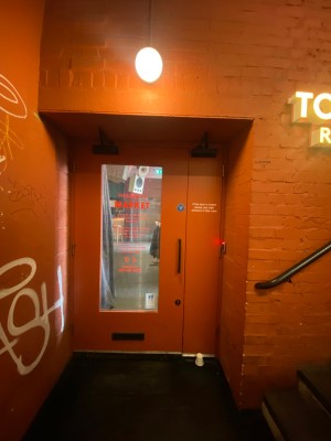 Red glass panelled door with red doorframe set back from the entrance. Black handle on the right. Pull to open.