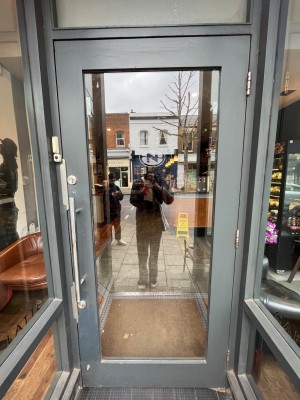 Grey framed glass door. Pull open via silver metal handle on left. Doorbell on left side of doorframe.