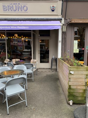 From sidewalk facing venue. Tables and chairs along left. Planter decor along right. Path down centre to main entrance.
