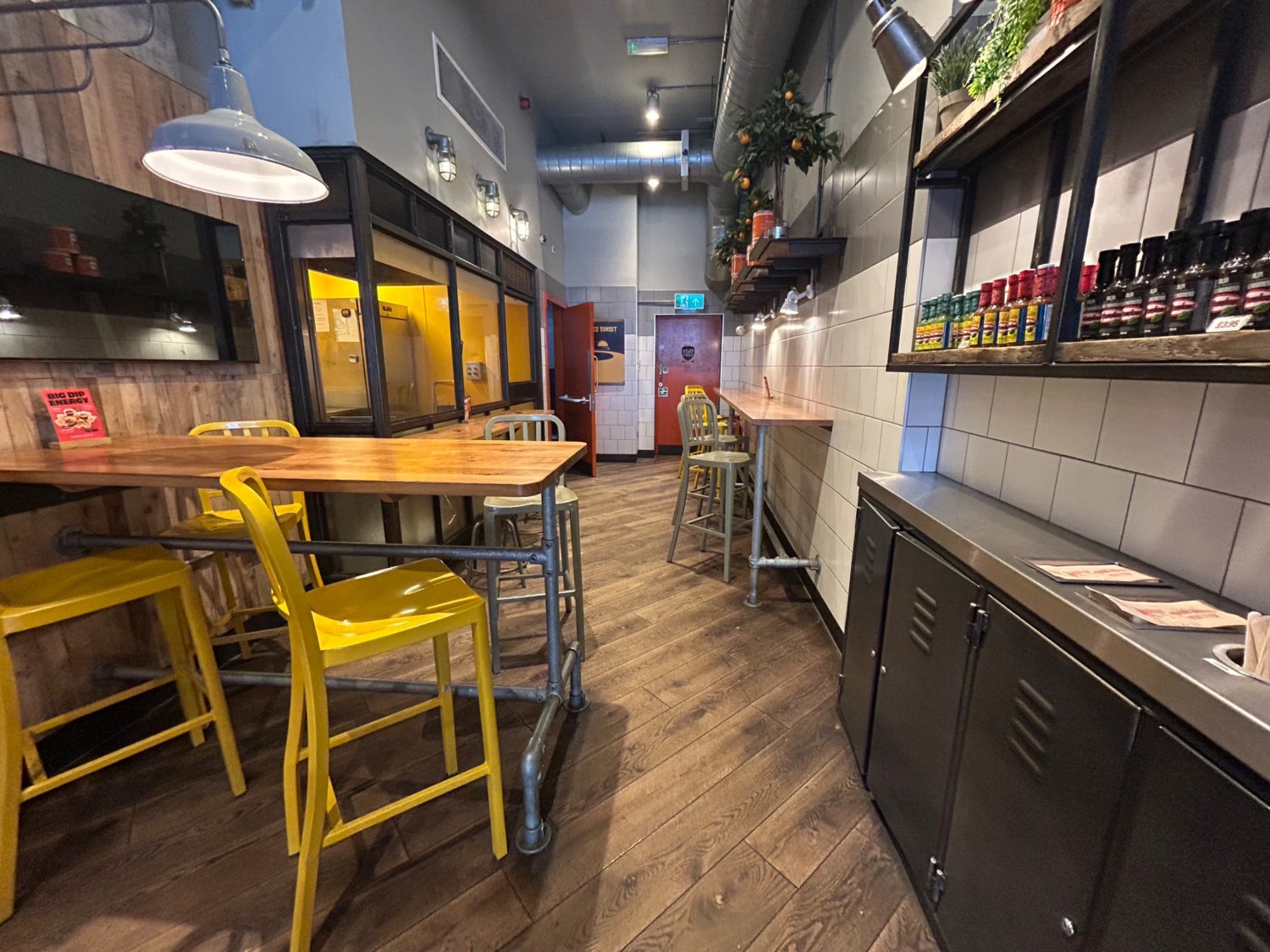 From further along pathway, facing straight. High tables and stools on left, and further ahead on right wall.