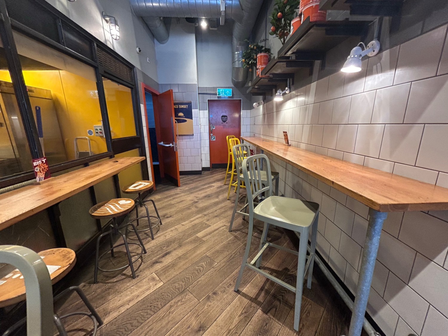 From end of high tables on left, facing straight. High tables and stools along left and right walls. Path in middle leads to bathroom on back left.