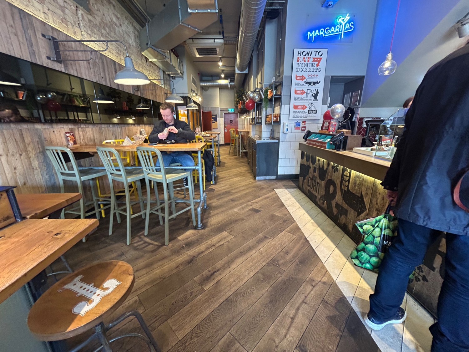 From end of high table and stools, facing straight. Counter along right wall. High tables and stools along left side. Path directly ahead.