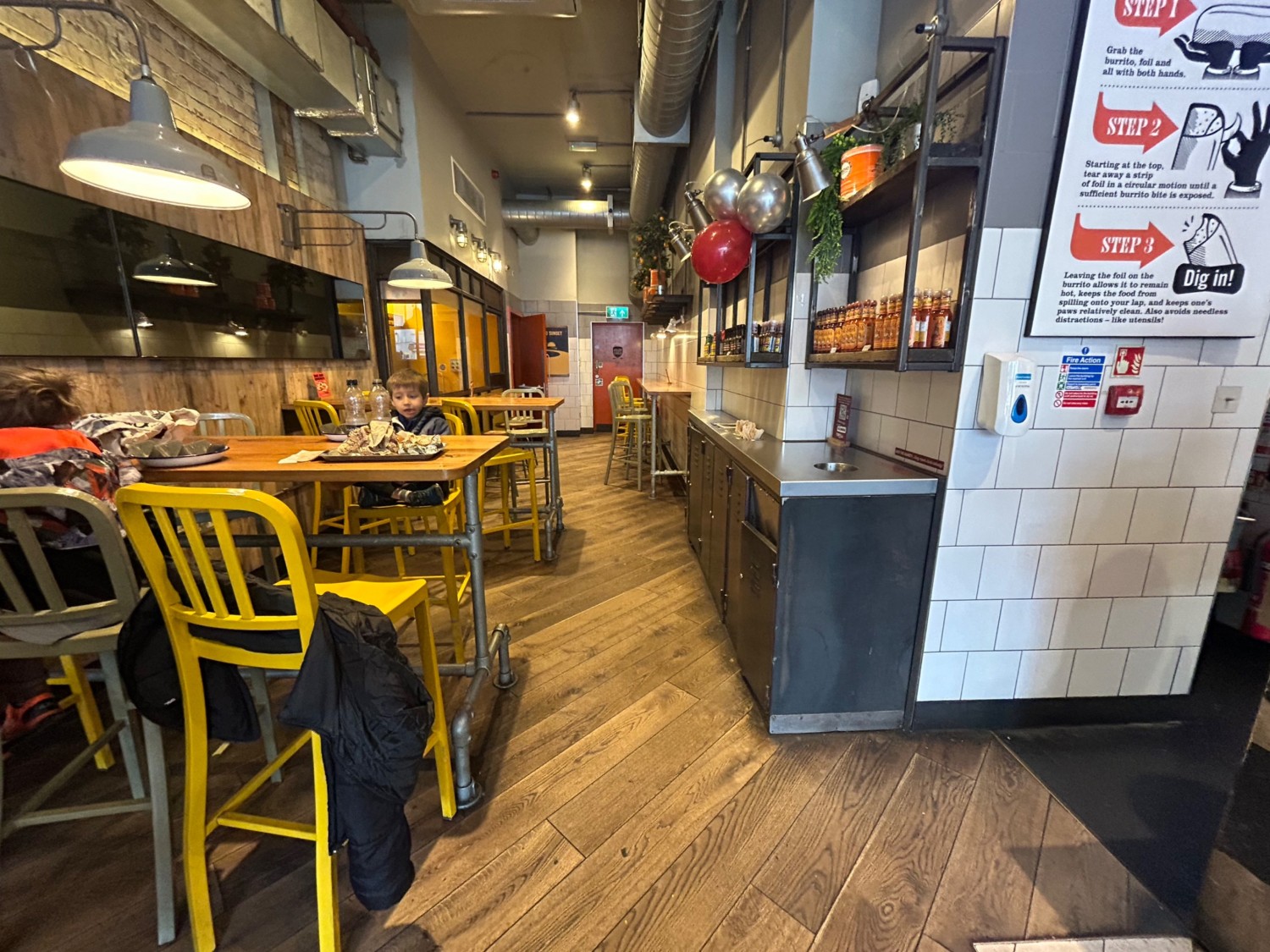 From end of counter, facing straight. High tables and stools continue along left side. Path straight ahead.