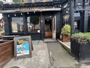 From pavement facing venue. Wooden divider, A-frame and picnic bench on left. Planters on right. Entrance at end of path ahead.