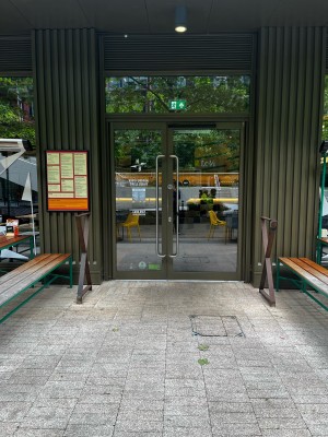 From sidewalk facing venue. Picnic bench and divider on left. Entrance ahead. Divider and picnic bench on right.