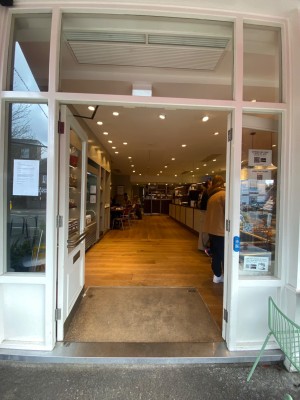 Double glass door, with white wooden doorframe. Doorbell for assistance on right. Horizontal golden handles at centre of doors. Push to open.