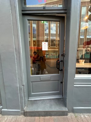 Light grey framed door with large glass panel. Black vertical handle on right. Push to open.