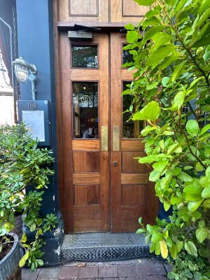Double brown wooden doors with glass panels. Push from middle to open. Plants on both sides of door. Right slightly obstructed by plant.