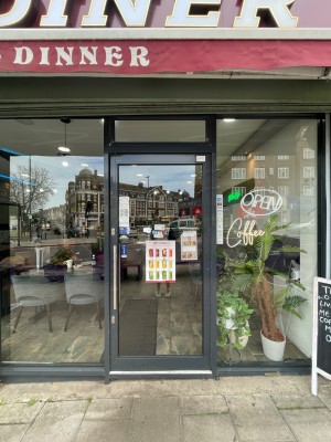 Black glass door, with handle on left. Push to open. Outdoor seating at left and display board at right of entrance.
