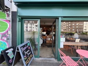Single glass door with dark green metal frame. Push metal handle on left to open. Sandwich board left. Table and chairs right.