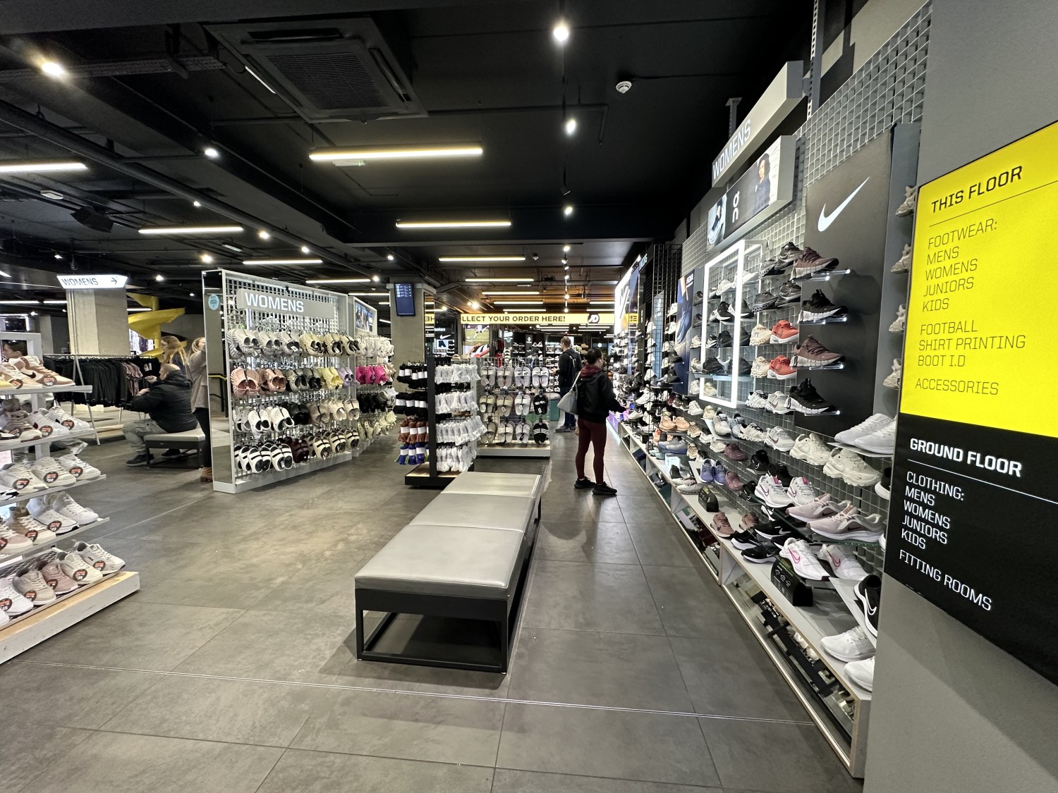 From lift looking right, shoes displays on left side with wide path straight ahead. Benches in centre of space to left of path, display stands behind.