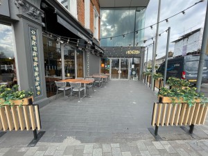 From sidewalk facing venue. Path to entrance proceeds between fences ahead. Outdoor seating beyond fences to left. Path has upwards slope.