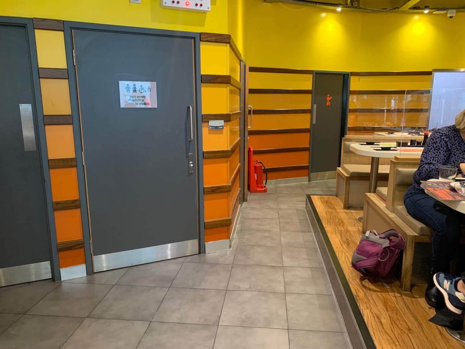 From raised booth seating facing left. Booths along right side. Toilet doors along left. Pathway does centre veers right along back wall.