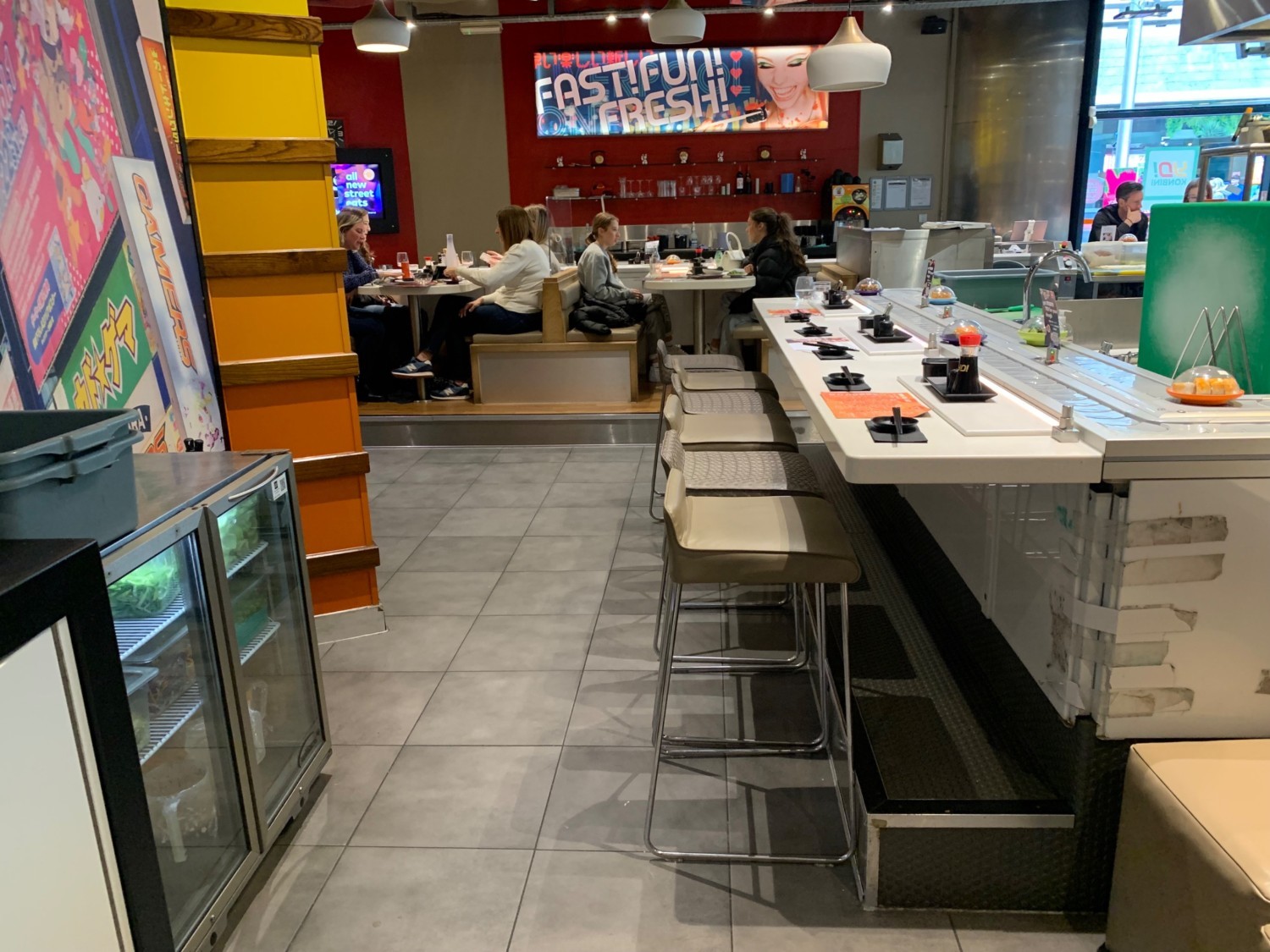 From end of booths facing right. Fridge to left. High tables and stools along right. Pathway down centre to raised booth seating.