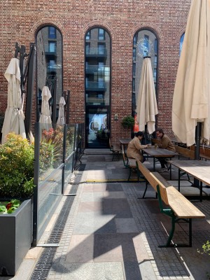 From sidewalk facing venue. Fence along left. Outdoor seating along right. Pathway down centre to main entrance.