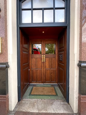From pavement looking straight at open archway followed by wood double doors with glass panels. Gold handles towards top centre of door. Pull to open.