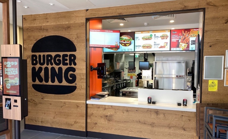 A small venue. 3 self service touch screens are found to the left of a central counter. A wall mounted screen shows order status.