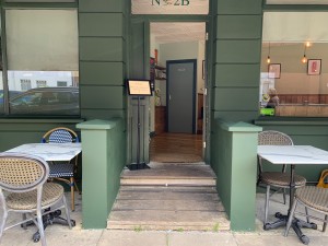 From street, facing venue. Path ahead with dividers on both sides to green wooden door. Push from left to open. Outdoor seating beside dividers.