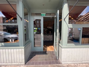 Double glass doors in blue wooden frame set back from pavement. Pull silver metal handle from centre to open.