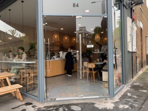 From street, facing entrance. Outdoor seating to left of entrance. Path straight to entrance doors ahead.