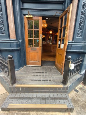Facing entrance door. Brown framed door. Pull golden handle in centre to open. Black fence on left & right of door. Assistance bell on right fence.
