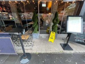 From sidewalk facing venue. Portable signs on right. Fence on left. Seating area behind on left. Path to entrance straight ahead.