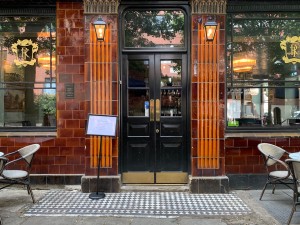 Black and glass panelled doors with gold push pads in centre of doors. A-frame to left before door. Tables and chairs to far left and right.