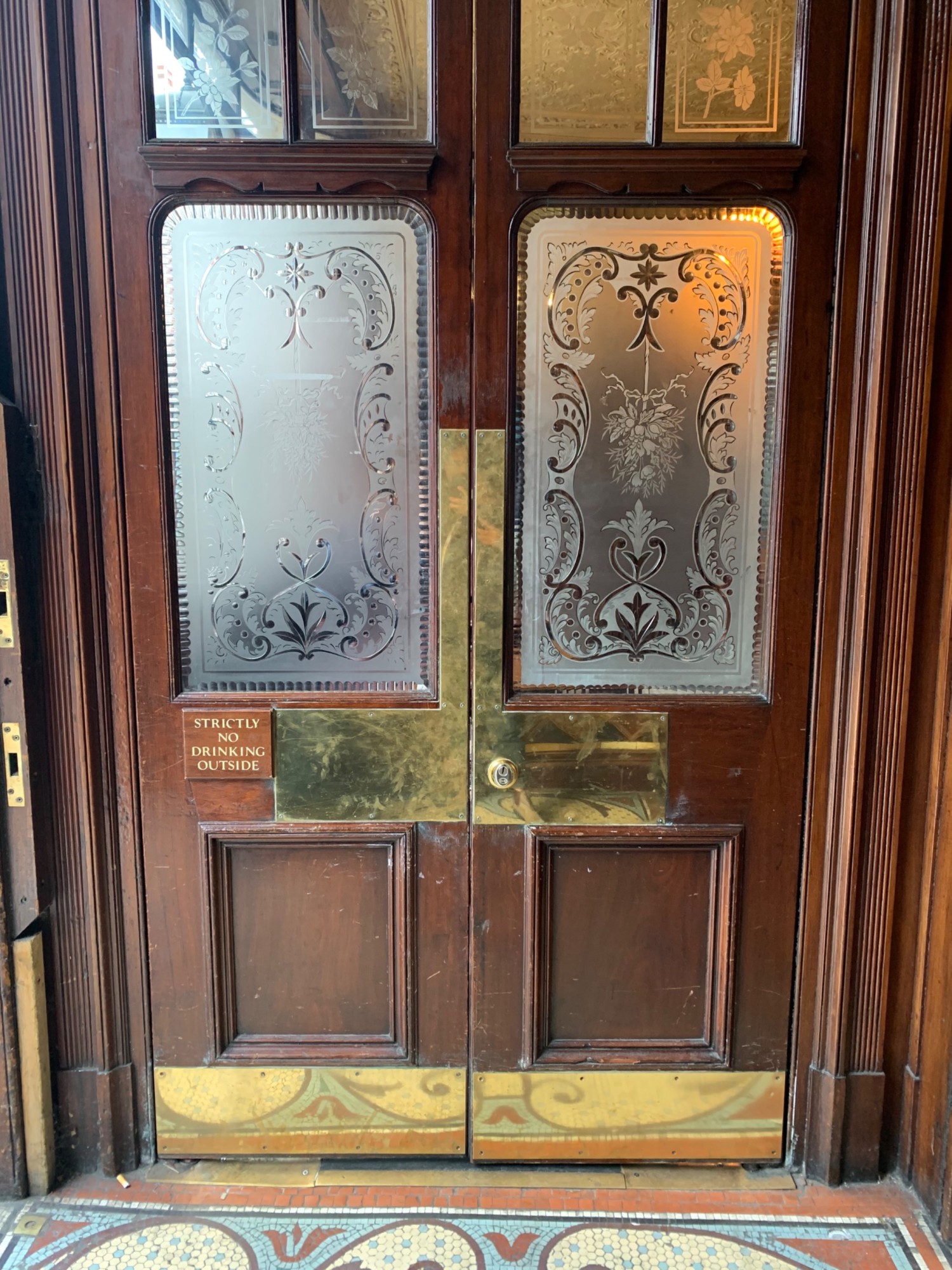 From right foyer, looking left. Doorway to main area ahead. Double brown wooden doors with glass panels. Push from center to open.