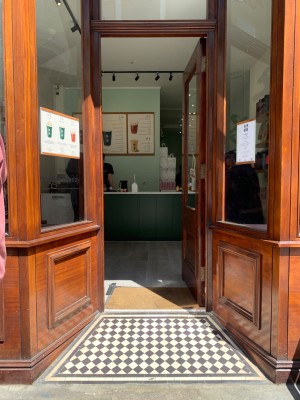 Single glass door with brown wooden frame. Push from gold metal handle on left of door to open.
