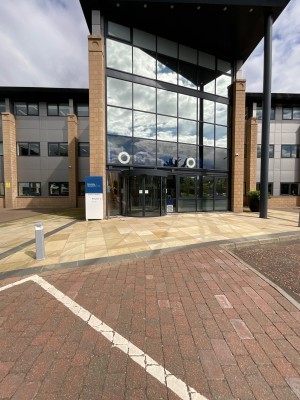 From car drop off space facing towards entrance. Pathway leads to glass double doors ahead. Intercom and swipe access to left of door.