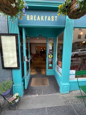 Glass door with blue frame. Handle at right, pull to open. Outdoor seating at right of entrance.