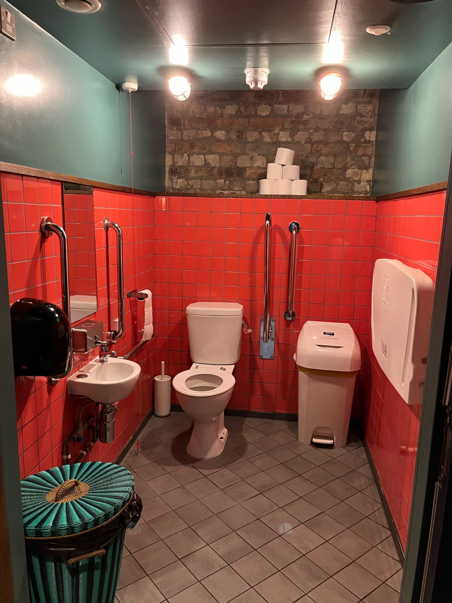 The interior of the accessible toilet. The toilet is at the back on the left, with the sink on the left wall. The bin is by the door on the left