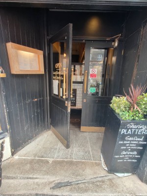 Facing venue.  Outdoor seating on immediate left. Path ahead to black framed entrance door. Pull golden metal handles in centre of doors to open.