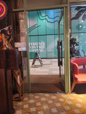 Doorway is wide with swing door propped open. There is an A Frame sign outside in the middle of a very wide pavement