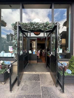 Double black wooden doors with glass panels. Gold metal handles in centre, pull to open. Dividers left and right. Push button to left of doors.