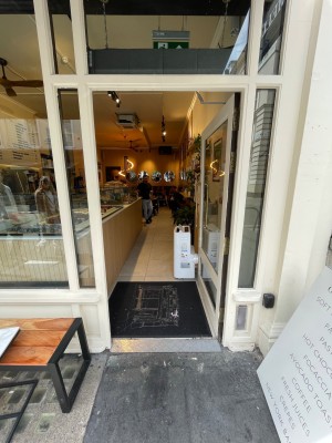 White glass door with handle at left, push to open. Display board at right, outdoor seating at left.