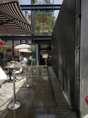 From sidewalk facing venue. Wall fountain along the right. Path to entrance ahead. Rope barriers along the left. Seating behind.