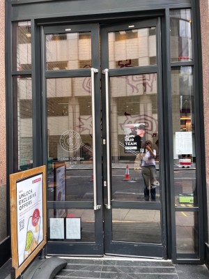 Glass doors with silver vertical handles in the Centre. Push to open. Standing sign in front of doors to left obstructing path.