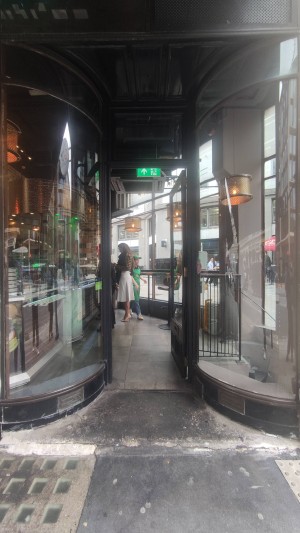 Glass door with wooden frame ahead. Push door with vertical handle on left. Door fixed open with a chain.