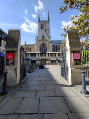 From pavement looking straight. Black metal gates ahead, push from centre to open. Bollards in front of gates to left and right. Planter beyond gates.