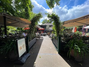 From parc facing venue. Green metal gate ahead. Push on left to open. Plants either side. Path to entrance straight ahead through outdoor area.